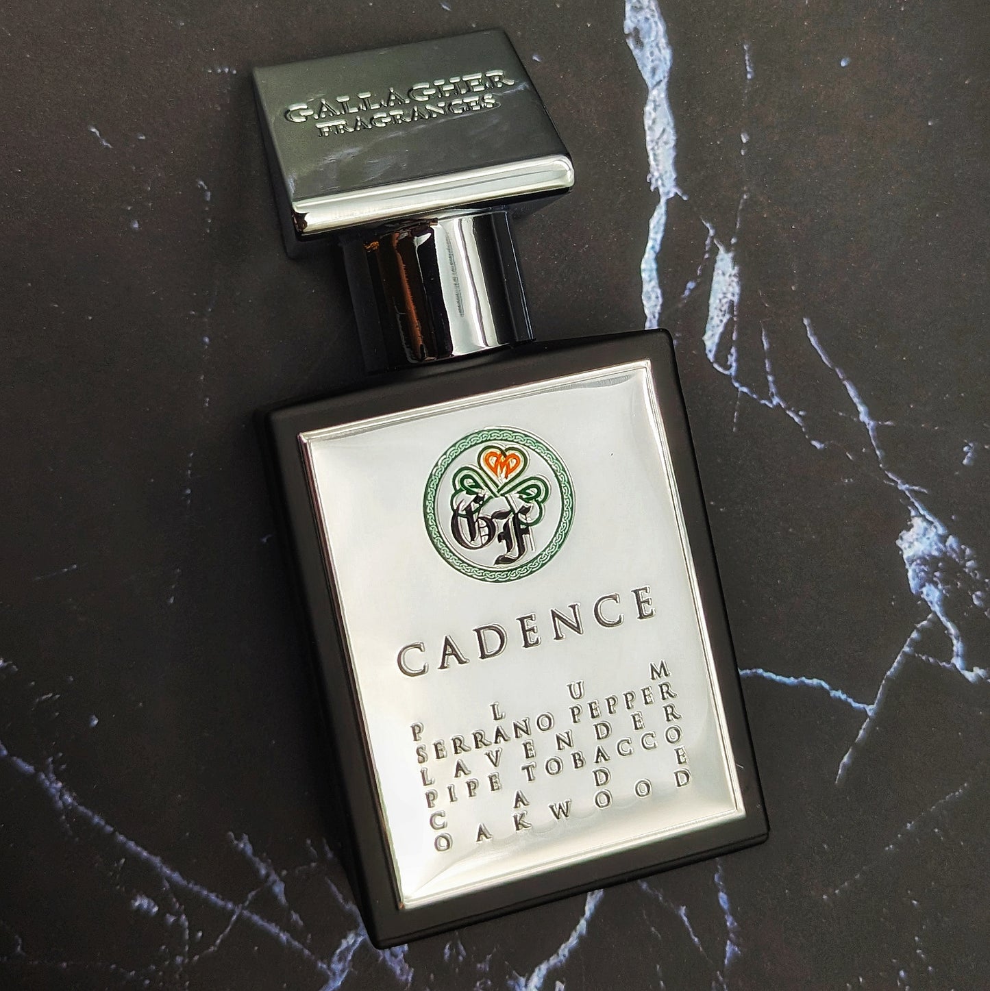 A bottle of 'Cadence' fragrance with a label that lists the key ingredients such as Plum, Serrano Pepper, Lavender, Cinnamon, Pipe Tobacco, and Oakwood.