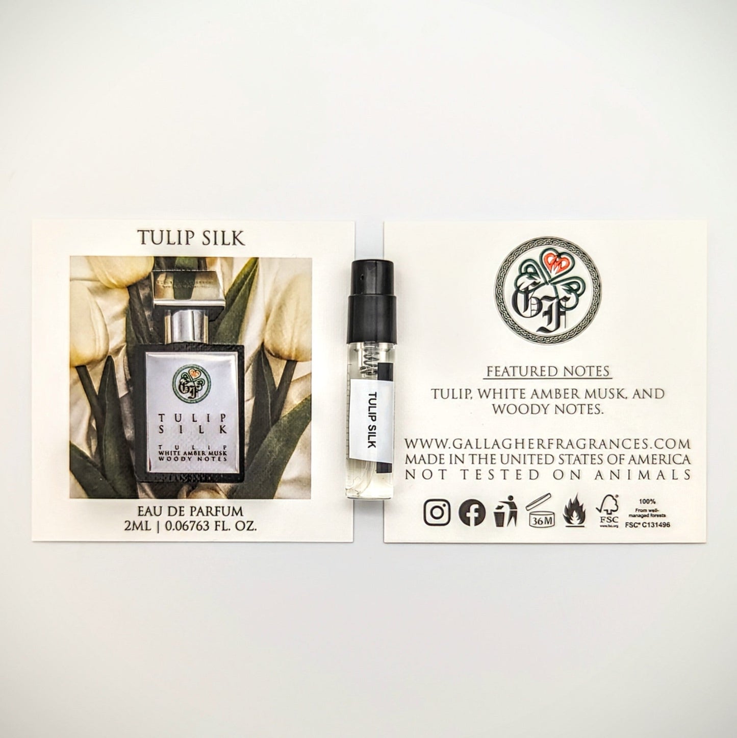 A bottle of Tulip Silk perfume placed among white tulips with a label that reads 'TULIP SILK.' With Featured Notes of Tulip, White Amber Musk, and Woody Notes. The background is white and appears to be a close-up shot of the bottle and flowers.