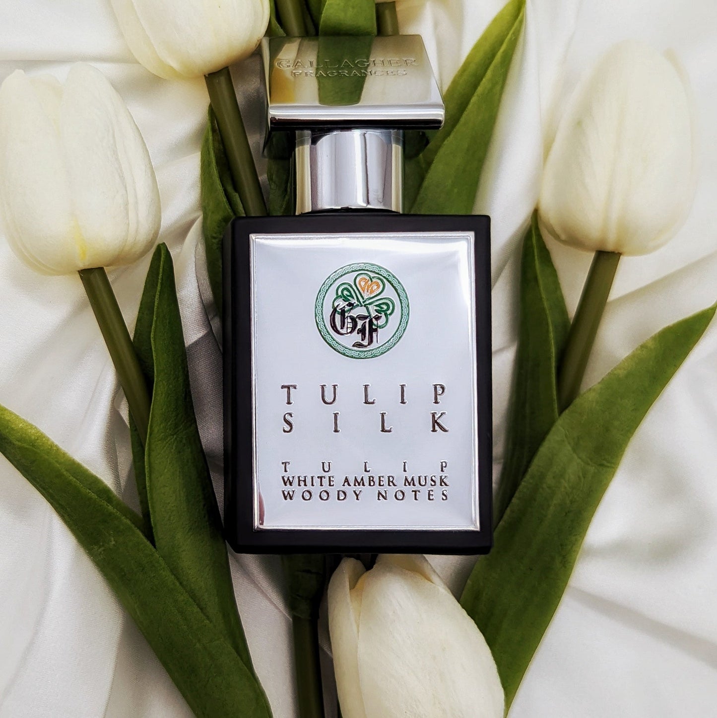 A bottle of Tulip Silk perfume placed among white tulips with a label that reads Featured Notes of Tulip, White Amber Musk, and Woody Notes. The background is white and appears to be a close-up shot of the bottle and flowers.
